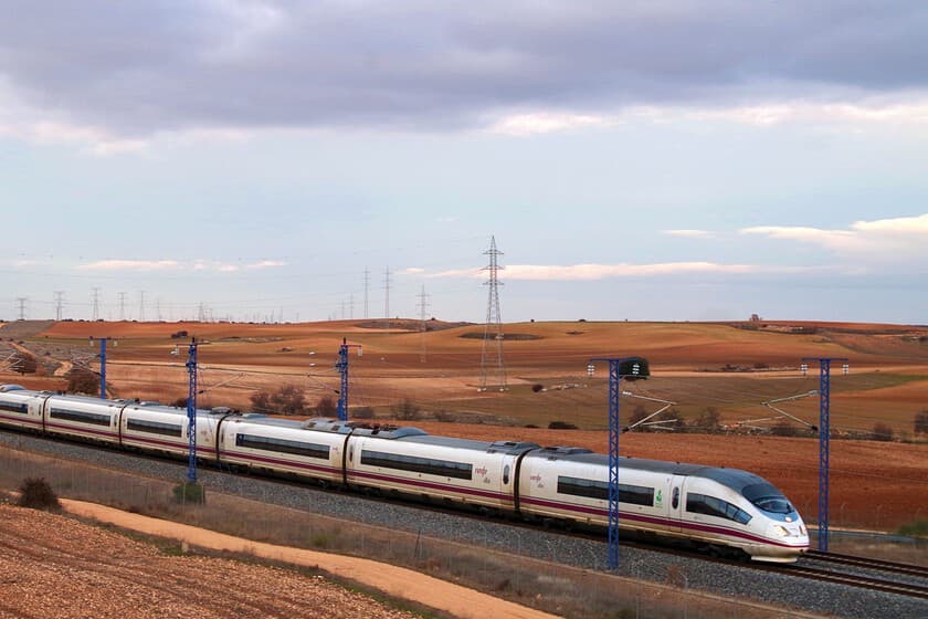 Tren de alta velocidad cruzando un paisaje rural al amanecer, con reflejos naranjas en los vagones.