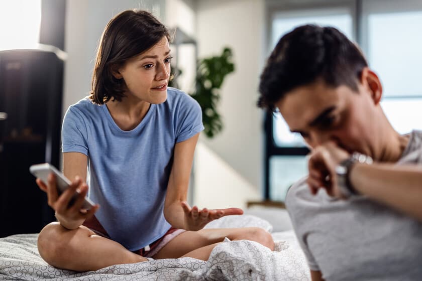 Persona mirando su teléfono mientras está sentada en una cama junto a su pareja que duerme, luz tenue