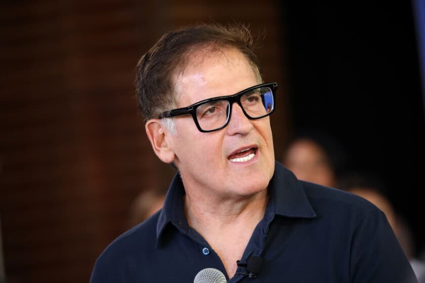 Mark Cuban hablando en un evento tecnológico, con fondo de pantalla de código de IA