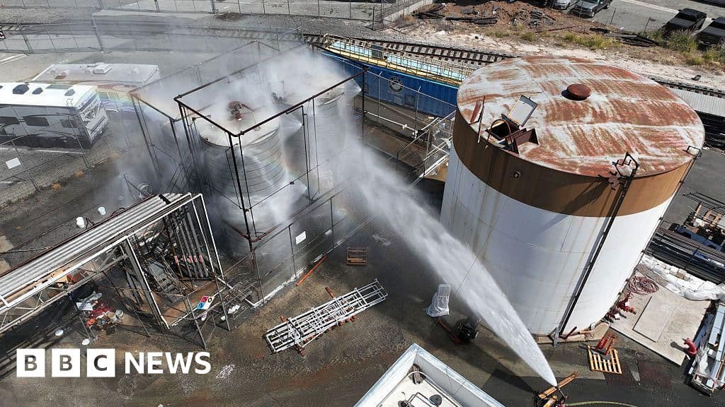Bomberos con trajes hazmat cerca de un contenedor industrial con humo naranja
