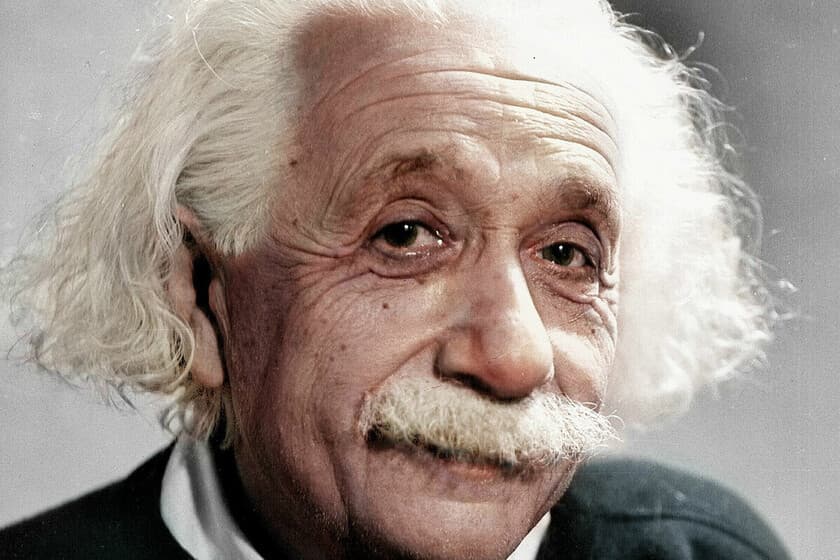 Fotografía de Albert Einstein en su escritorio, mirando pensativo, con fondo de pizarra con ecuaciones