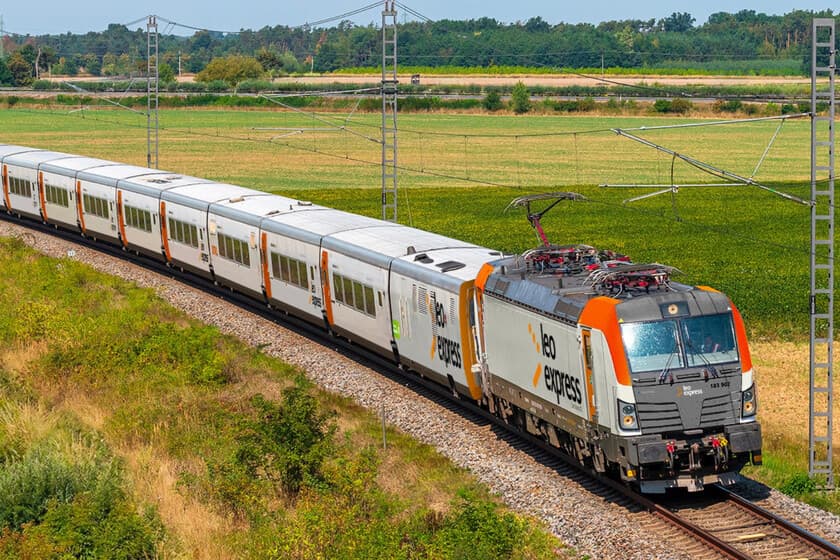 Tren moderno cruzando un paisaje europeo con vías y campos verdes