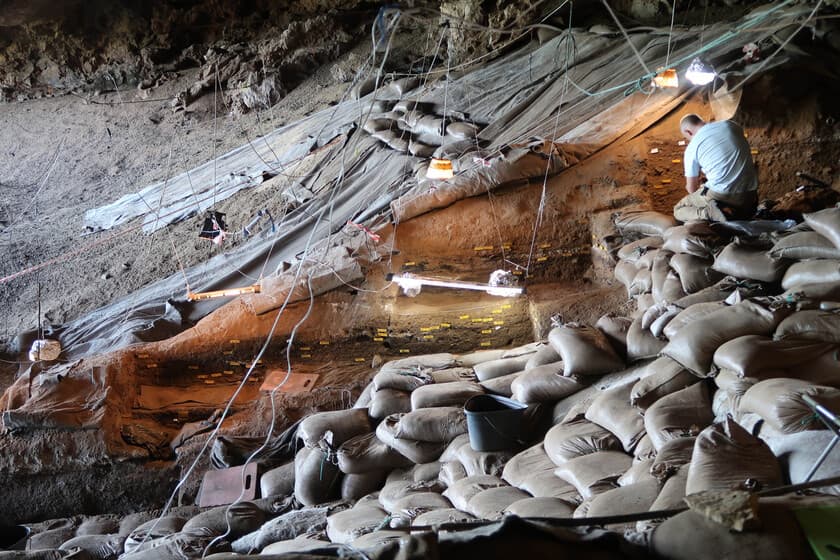 Representación artística de humanos prehistóricos acostados sobre camas de hierba en una cueva iluminada por una hoguera